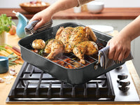 Roasted chicken being lifted from oven on a stove with veggies.