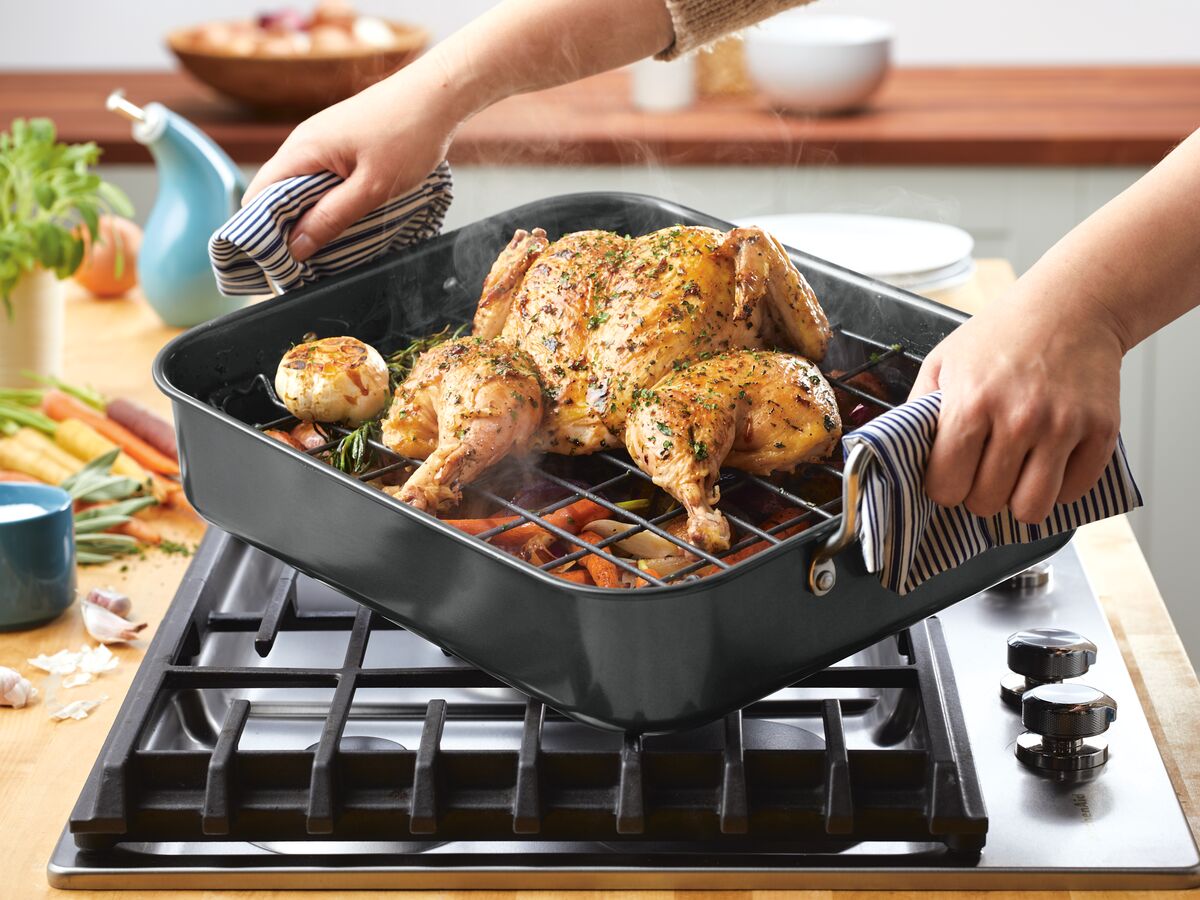 Roasted chicken being lifted from oven on a stove with veggies.