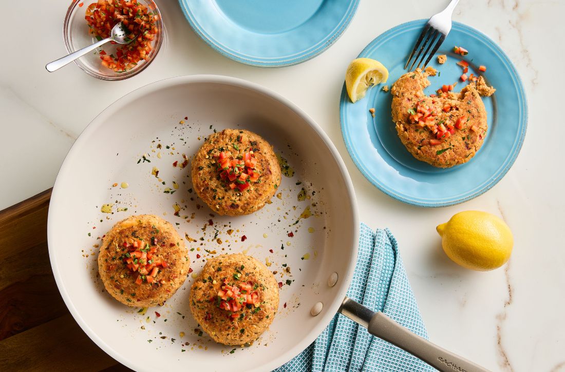 Salmon cakes being cooked in a Cucina Ceramic skillet in the shade Almond, with a finished salmon cake served on a light blue plate with lemon and garnish.