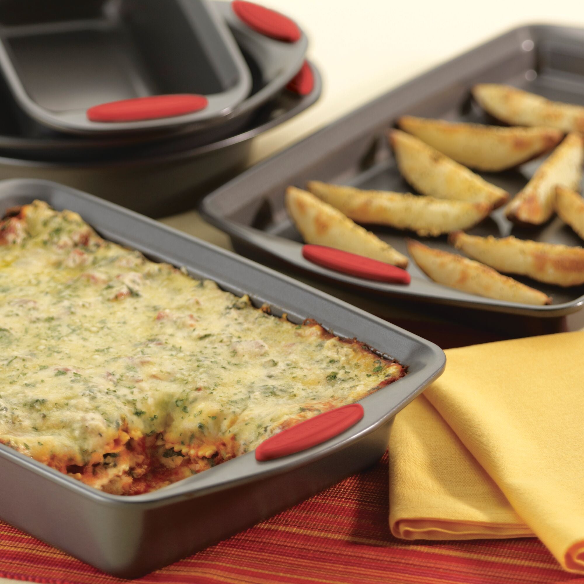 Baked lasagna and potato wedges in oven trays, with a yellow cloth nearby.