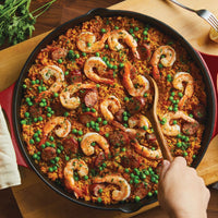 Stirring paella with shrimp, sausage, peas, and rice in a red-handled 14-Inch NITRO Cast Iron Skillet. This induction-ready pan by Rachael Ray is ideal for achieving the perfect simmer and is oven safe for applying finishing touches.