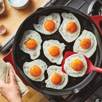 Seven fried eggs sizzle in a 14-Inch NITRO Cast Iron Skillet, with a spatula expertly lifting one. This induction-ready skillet, offered by Rachael Ray, is also oven safe, making it perfect for every culinary adventure.