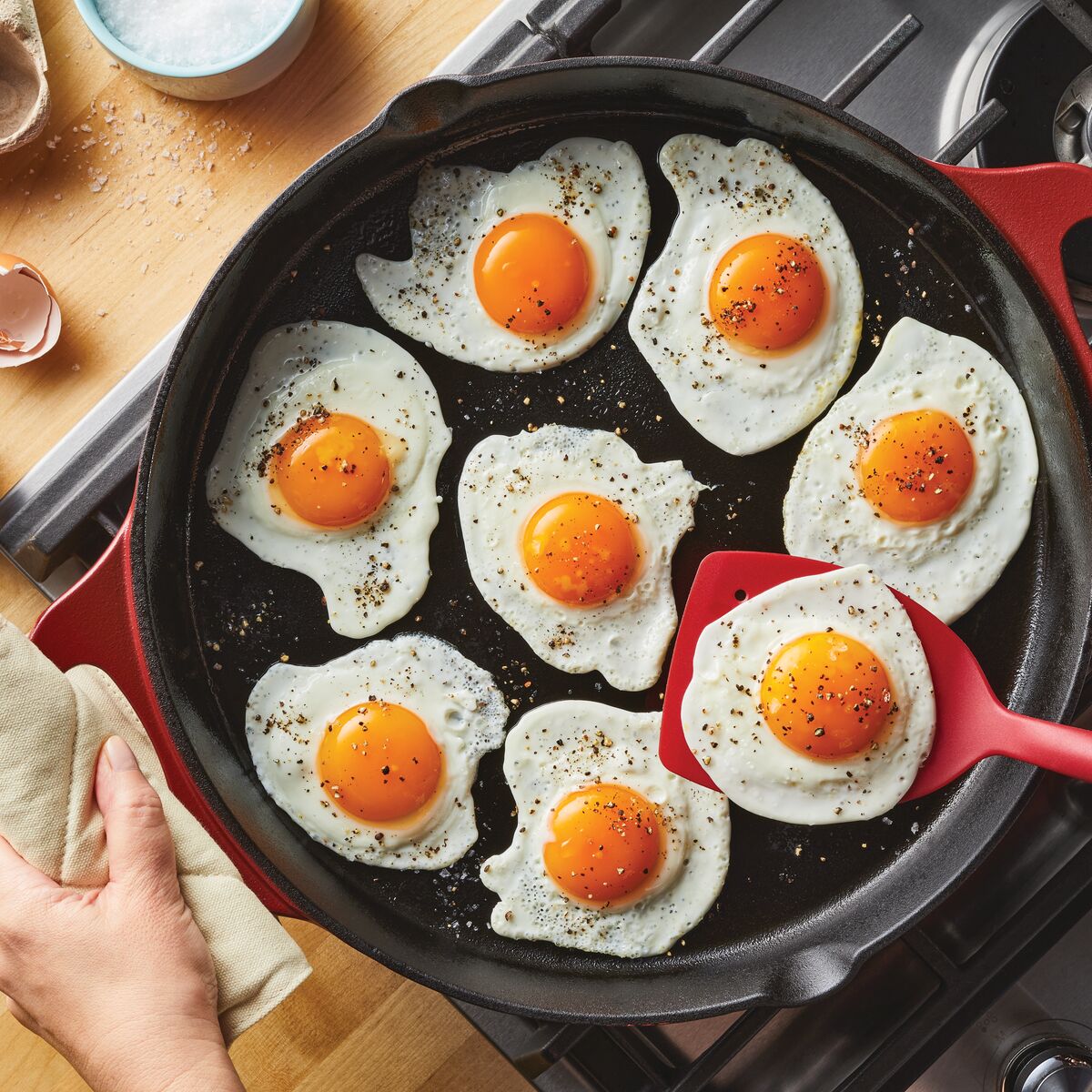 Seven fried eggs sizzle in a 14-Inch NITRO Cast Iron Skillet, with a spatula expertly lifting one. This induction-ready skillet, offered by Rachael Ray, is also oven safe, making it perfect for every culinary adventure.