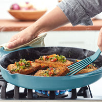 A person is cooking breaded patties with vegetables in a blue 14-Inch NITRO Cast Iron Skillet on an induction-ready stove.