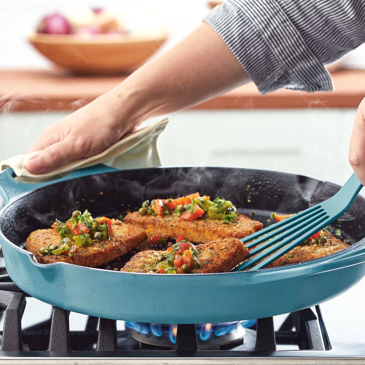 A person is cooking breaded patties with vegetables in a blue 14-Inch NITRO Cast Iron Skillet on an induction-ready stove.
