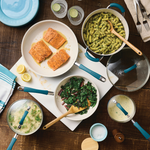 A tabletop with salmon, pasta, greens, and cookware sets.