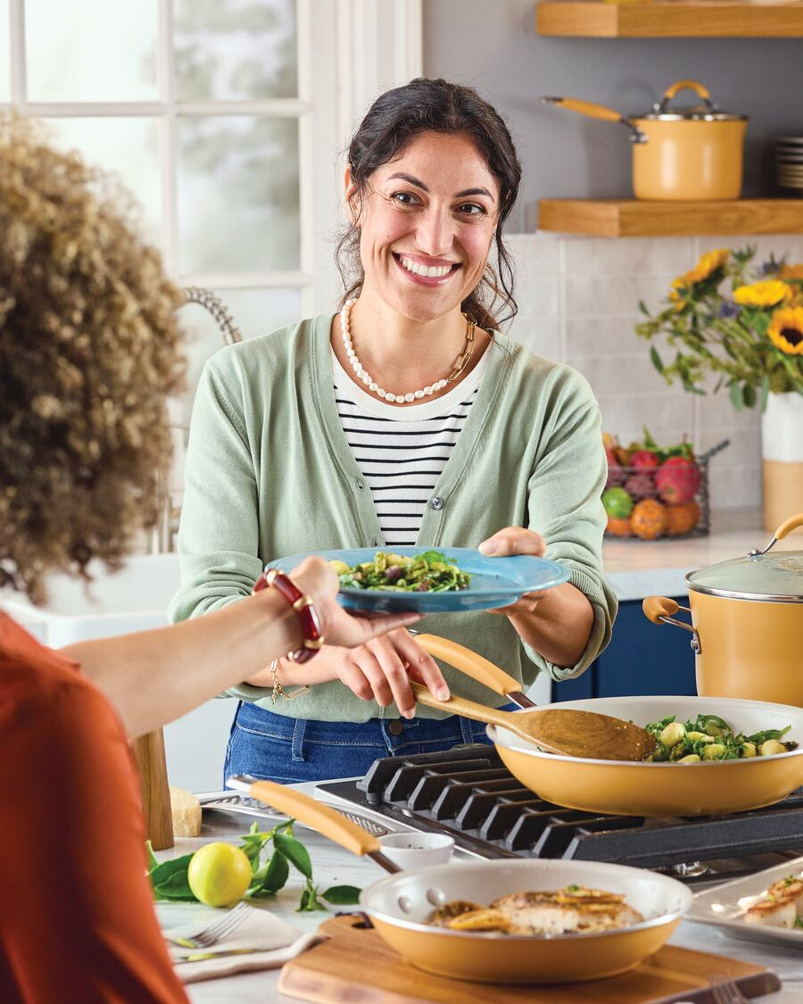 A woman smiling and serving a plate of food from a Rachael Ray Cucina Turmeric-colored Cucina Ceramic skillet in a bright, modern kitchen.