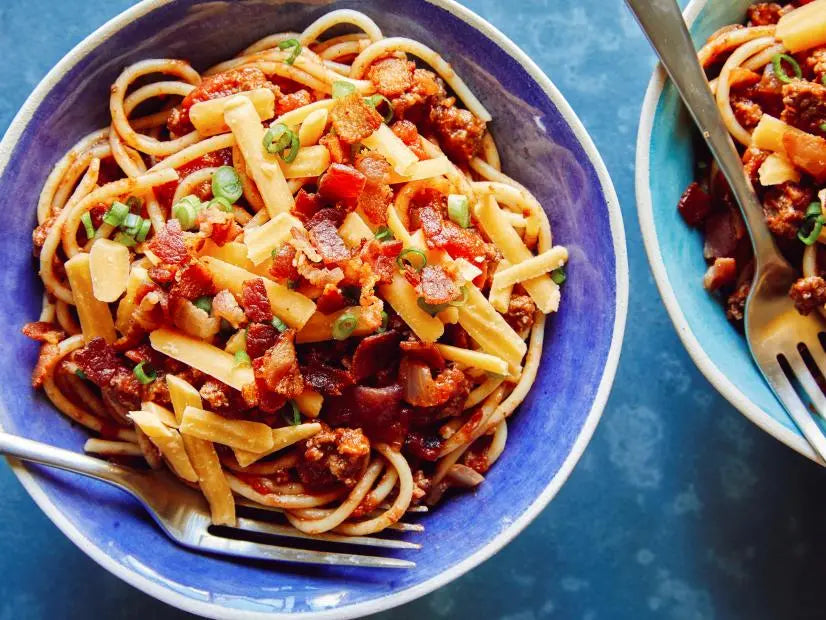 A blue bowl of Cowboy Spaghetti topped with crispy bacon bits, shredded cheese, and sliced green onions.