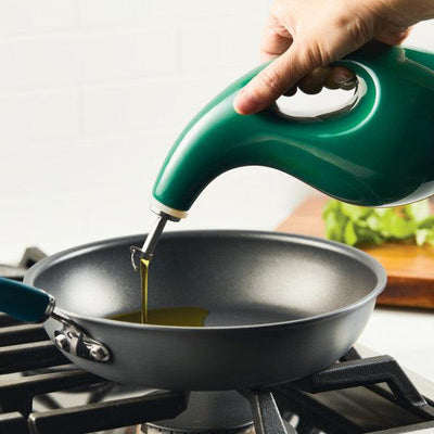 Pouring oil from a green dispenser into a skillet on a stove.