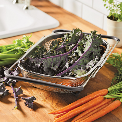 Colander with kale next to fresh carrots and green onions on a counter.