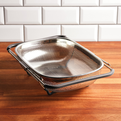 A metal mesh strainer with handles on a wooden kitchen counter.