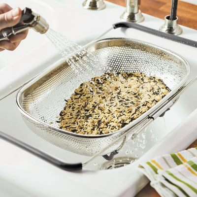 Rinsing rice in a mesh colander under faucet water in a kitchen sink.