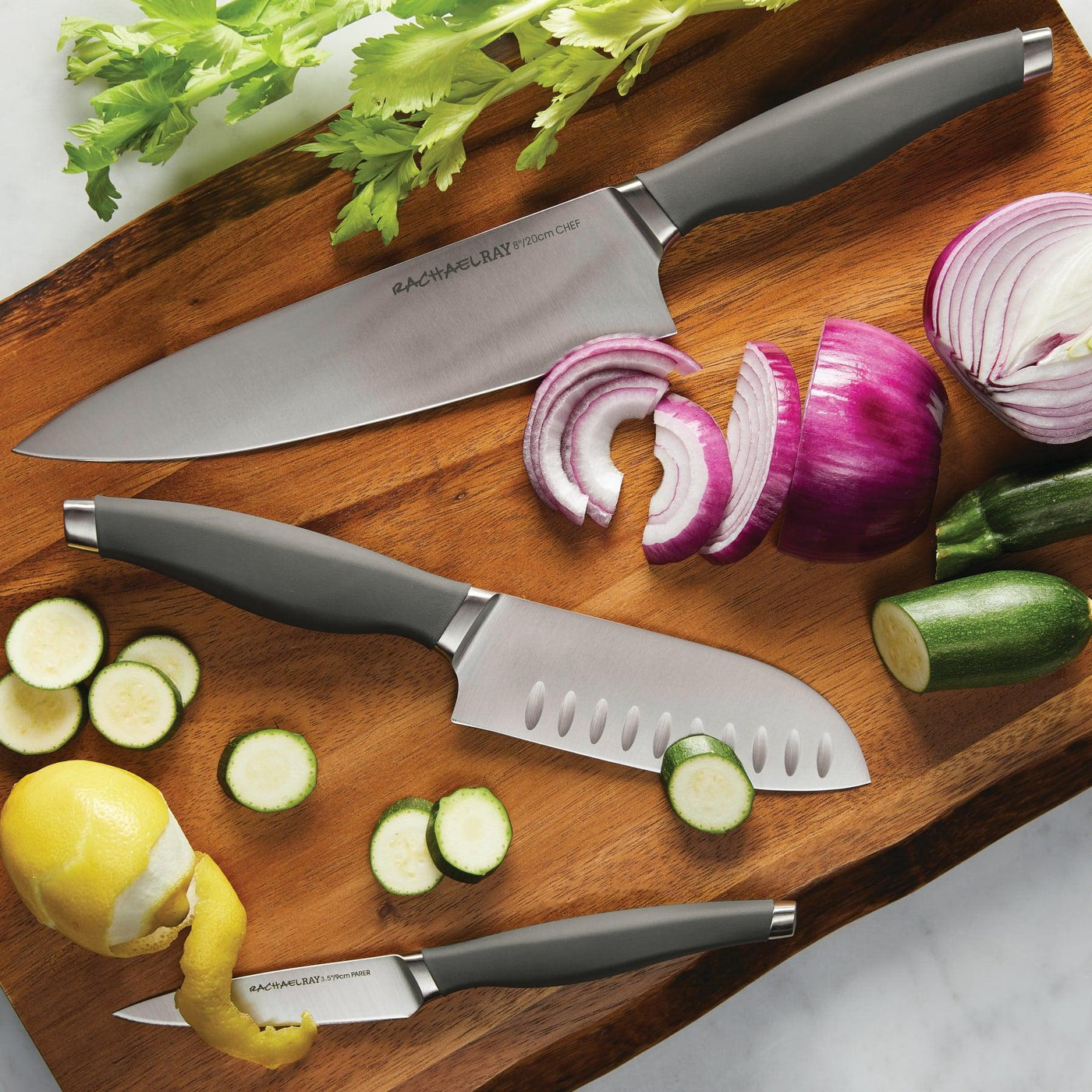 Three knives on a cutting board with zucchini, onion, and a peeled lemon.