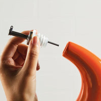 A hand holds the EVOO Bottle 24-Ounce stainless steel pour spout near an orange pitcher, poised for precision pouring.