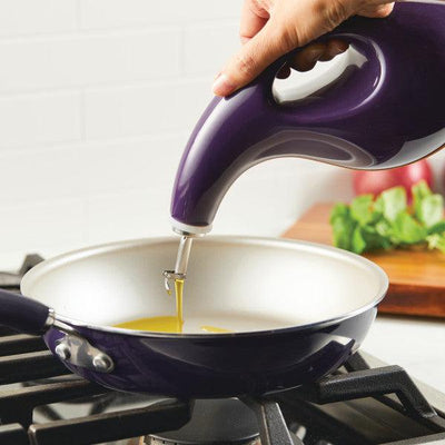 Hand pouring oil from a purple bottle into a frying pan on a stove.