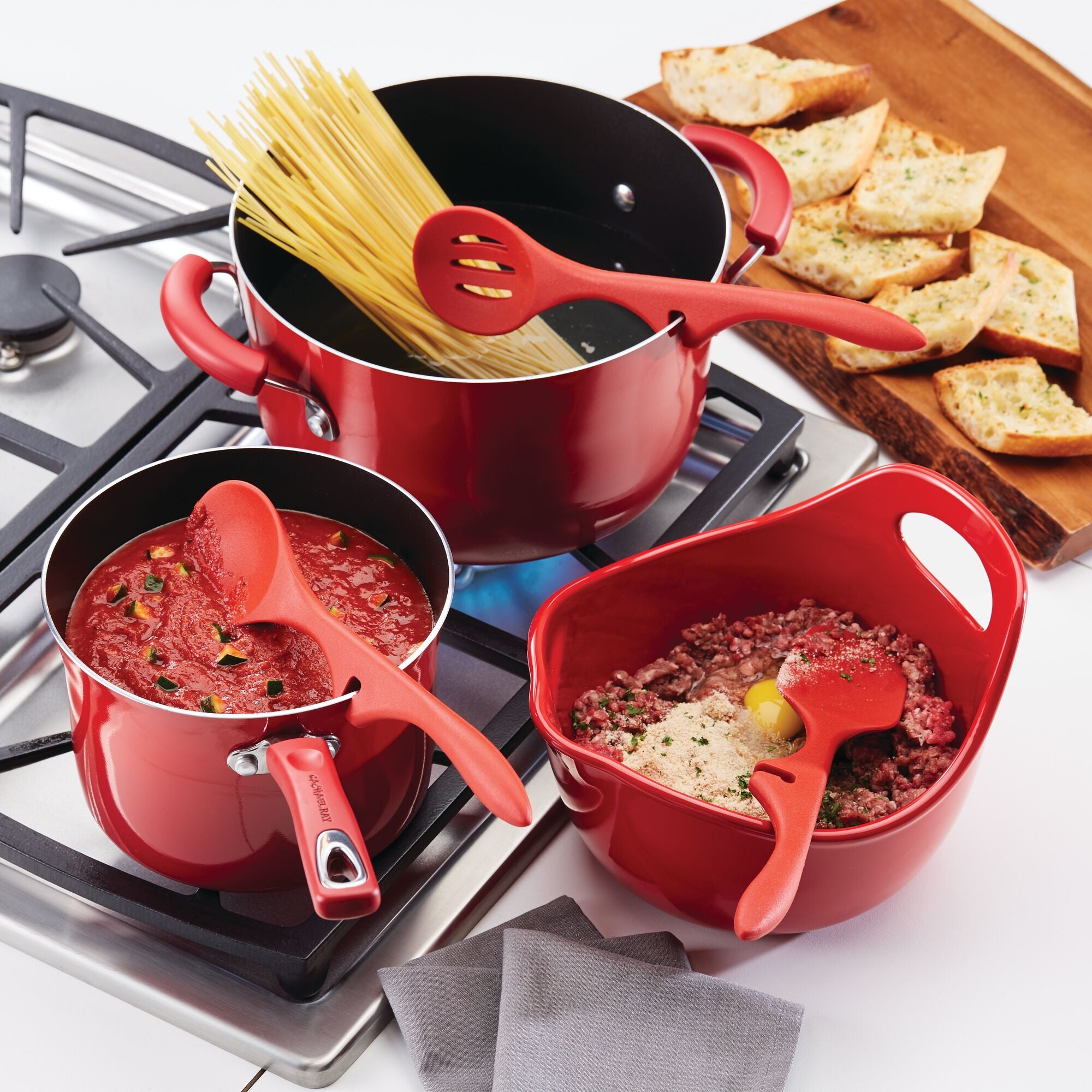 Red cookware with pasta, sauce, meat mixture, and garlic bread on a stove.