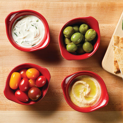 Four red bowls with dip, olives, cherry tomatoes, and hummus on a wooden table.