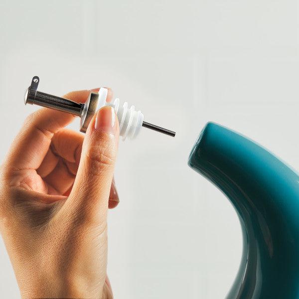 A hand grips the EVOO Bottle 24-Ounce beside a teal appliance spout, mastering the art of precision pouring with the finesse of a stainless steel pour spout.