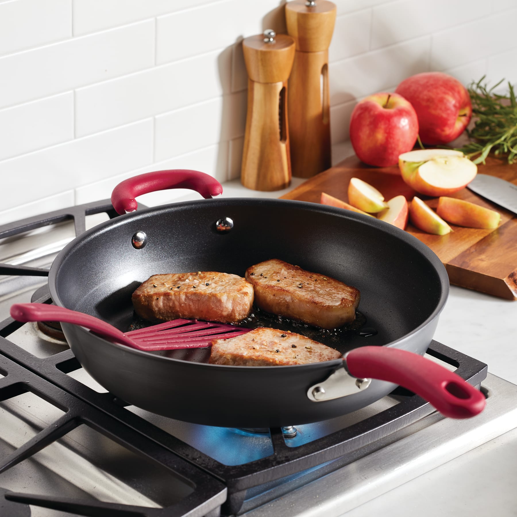 Three pork chops cooking in a pan on a stove with apples in the background.