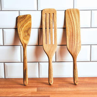 Three tools from the Acacia Wood Tool Sets are elegantly propped against a white tiled wall on a wooden countertop. These versatile spatulas, including a spurtle, are safe for use with nonstick cookware, ensuring your culinary creations remain pristine.