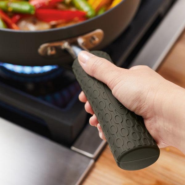 A person confidently grips the handle of a pan on the stove, where vibrant vegetables are cooking. The 3-Piece Silicone Sleeve Set offers a non-slip surface, ensuring a safe and comfortable hold, making the cooking experience both enjoyable and secure.