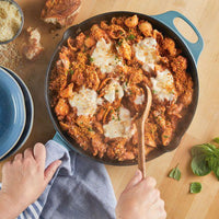 A person stirs baked pasta in a pre-seasoned NITRO cast iron skillet on a wooden table.
