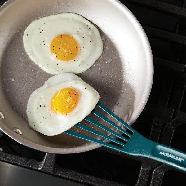 Two sunny-side-up eggs in a frying pan on a stove, with one being flipped with precision using the 10-Inch Nylon Spatula from the Nylon Spatula Set.