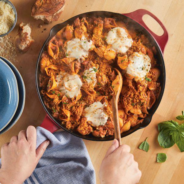 A person stirs a cheesy pasta dish in a 12-Inch NITRO Cast Iron Skillet on a wooden table.