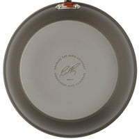 Bottom view of a round, gray pan from the 12-Piece Cucina Hard Anodized Nonstick Cookware Set, crafted from durable aluminum and featuring a label and signature.